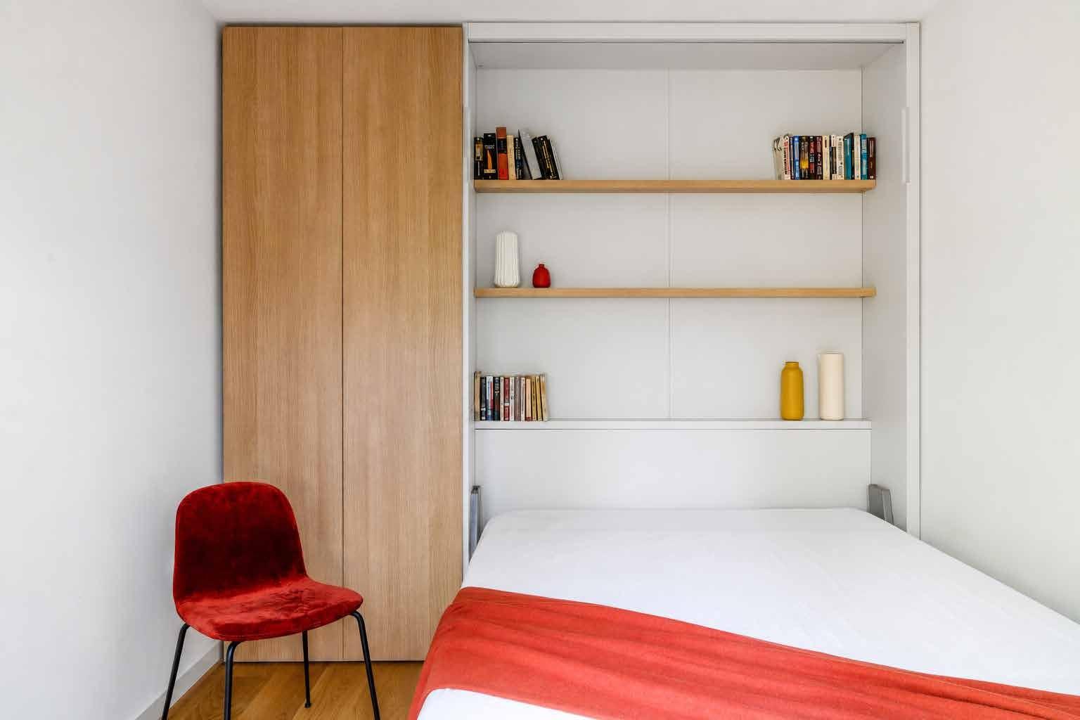 Fold-down bed in an oak wall unit with open shelves, a red chair beside it