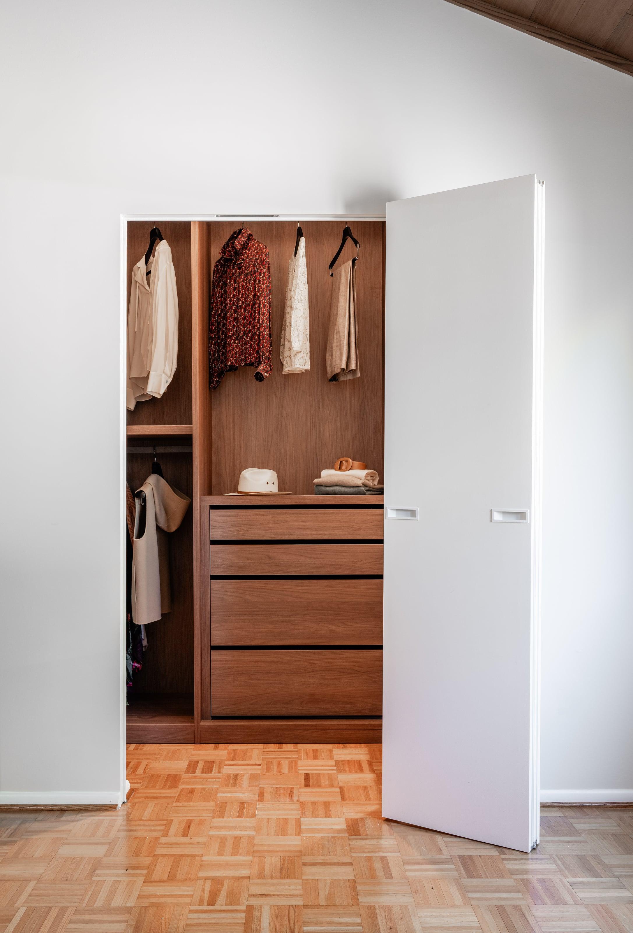 White pivot door open onto a walnut walk-in closet with drawers and hanging rails, on parquet