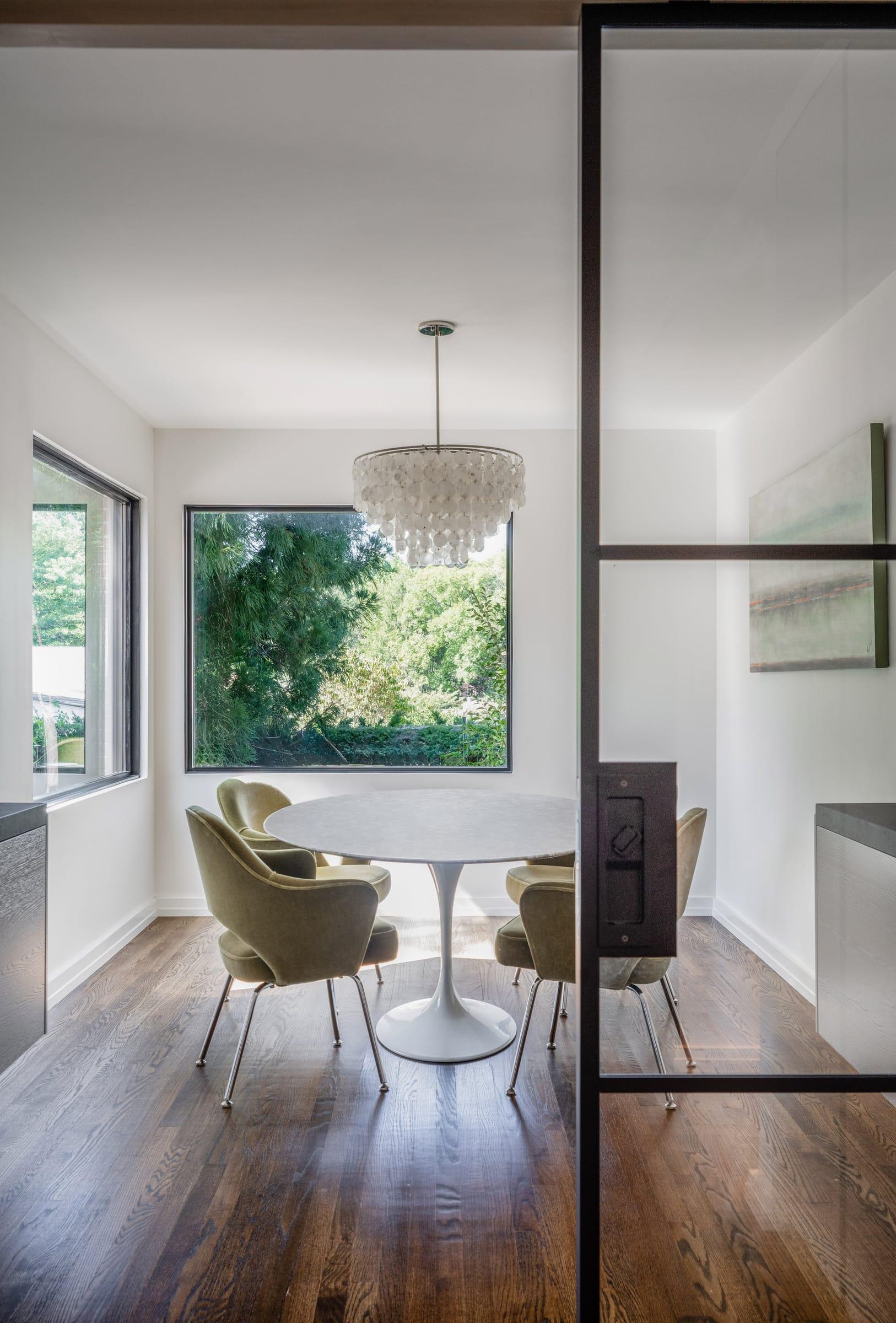 Round marble table with olive chairs under a shell chandelier, behind a black-framed glass door