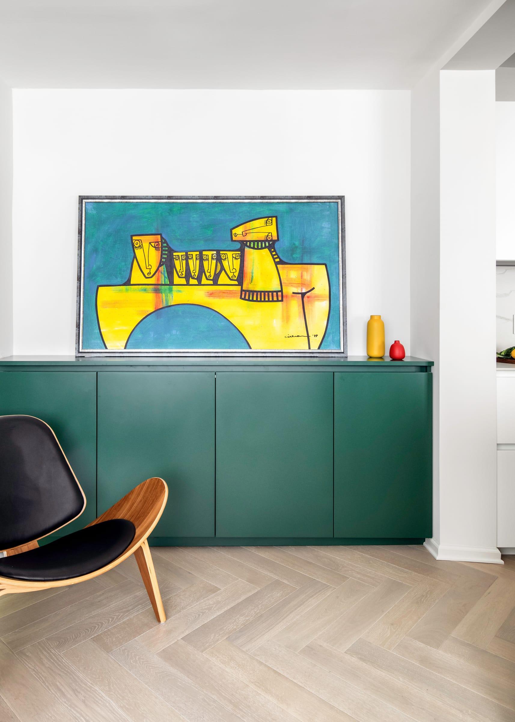 Green cabinets under a yellow painting, a wood lounge chair beside them