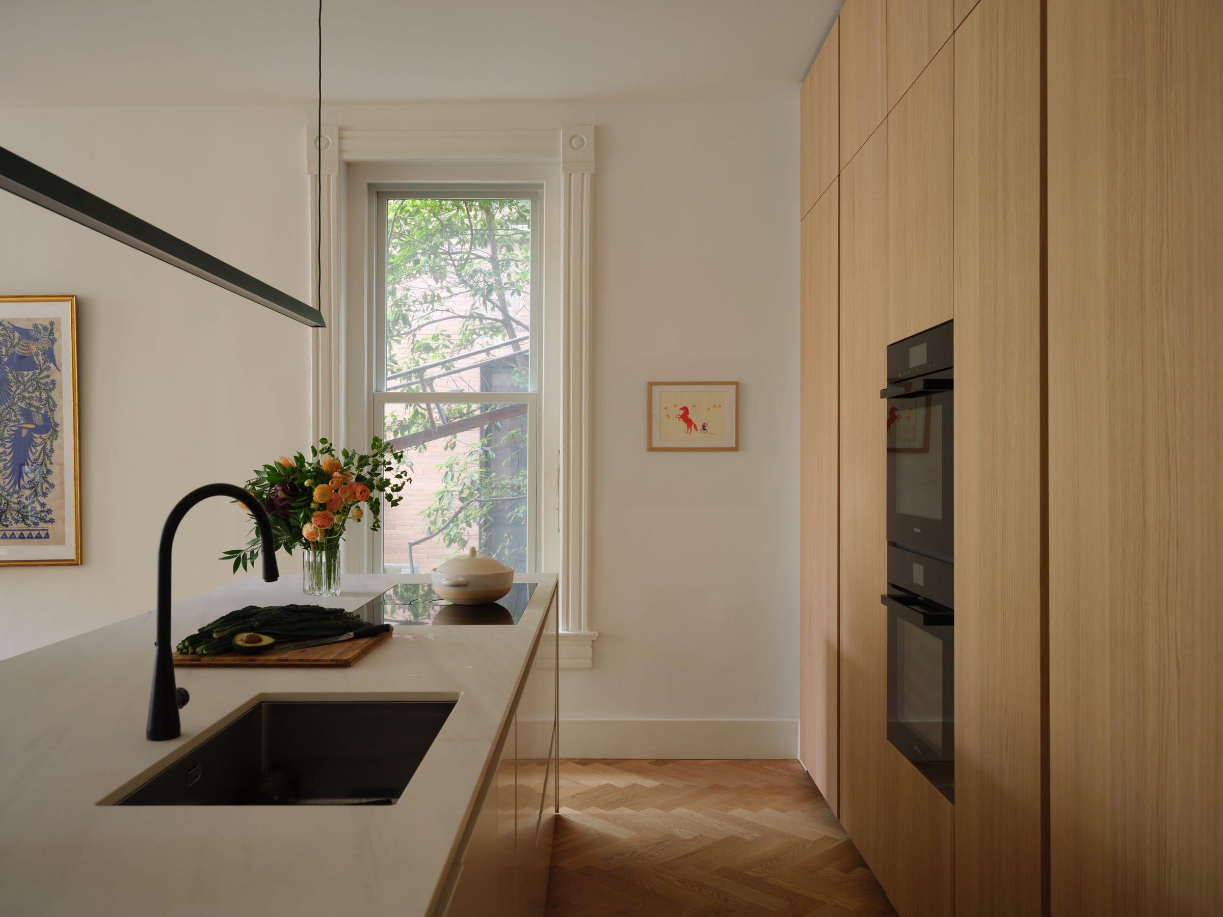 Kitchen in pale oak with a black tap and tall ovens, a painting by the window