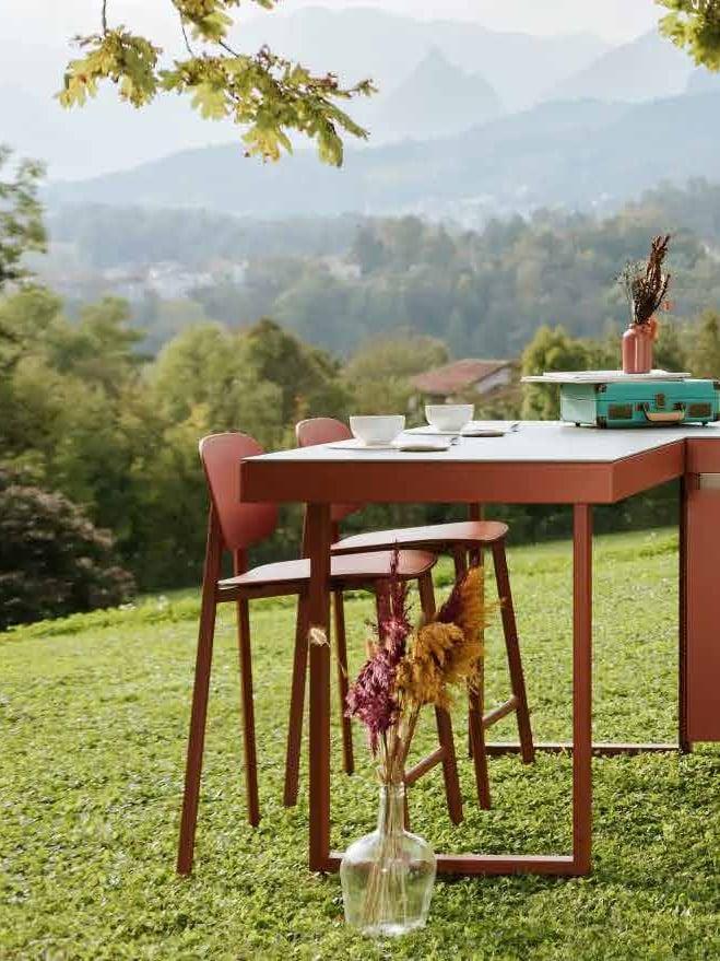 Rust-red outdoor kitchen with a bar table and stools on a lawn above a valley