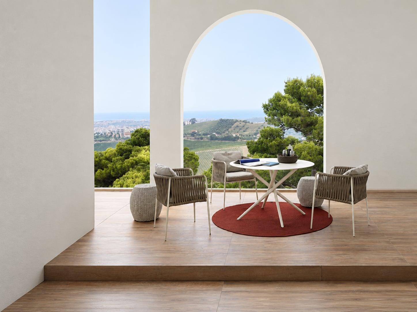 Outdoor dining table and armchairs on a red rug under an arch, looking out over the coast