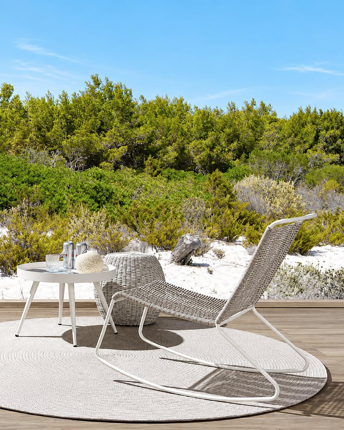 Woven white lounge chair and side table on a round rug on a dune